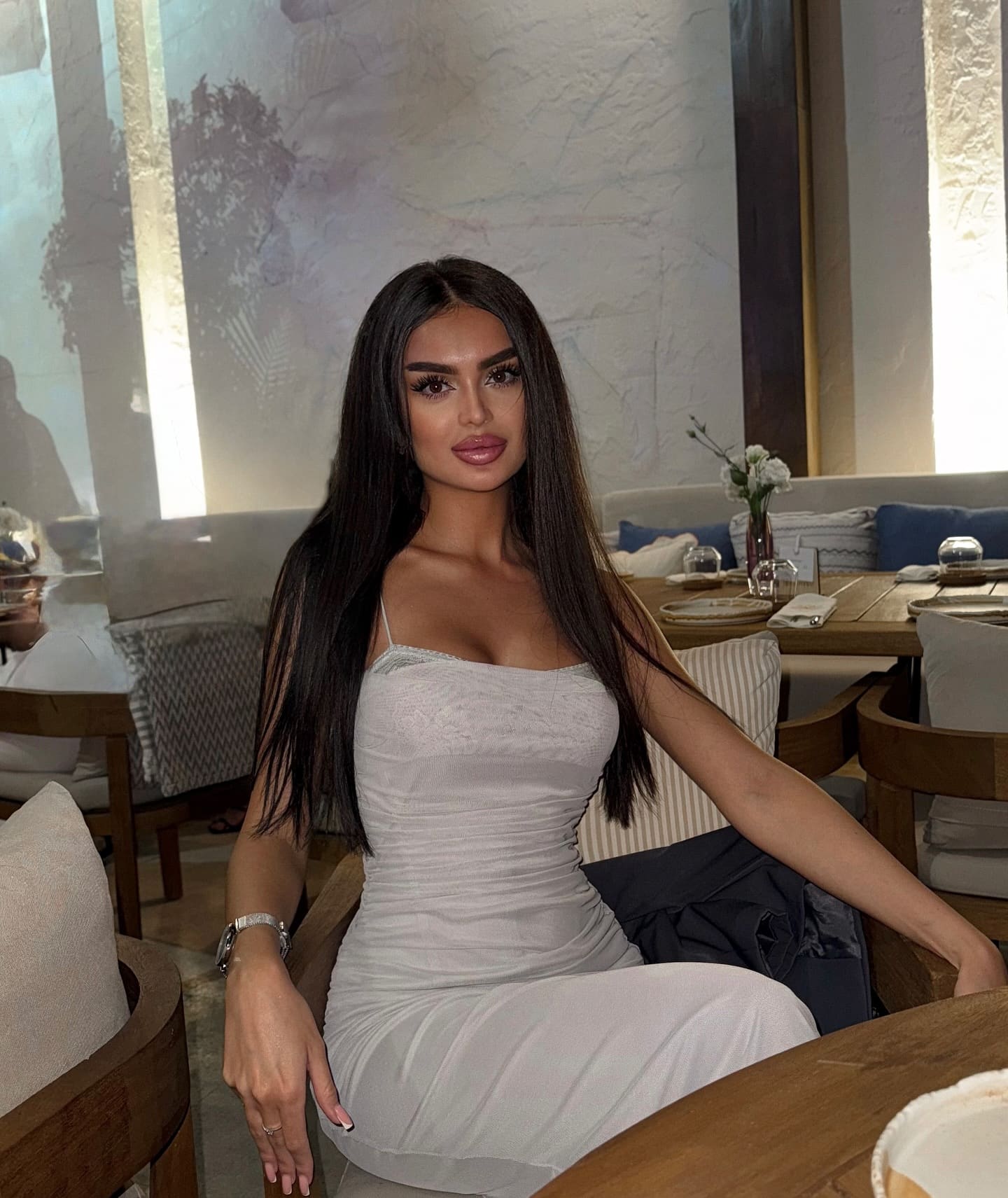 Sholpan sits in a wooden chair at a table, wearing a light-colored spaghetti-strap dress in a restaurant setting.