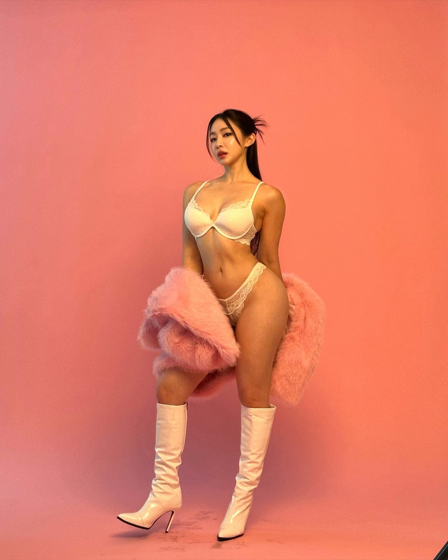 Against a pink background, Kali wears a white lace bra, high-cut panties, knee-high stiletto boots, and a pink fur coat.