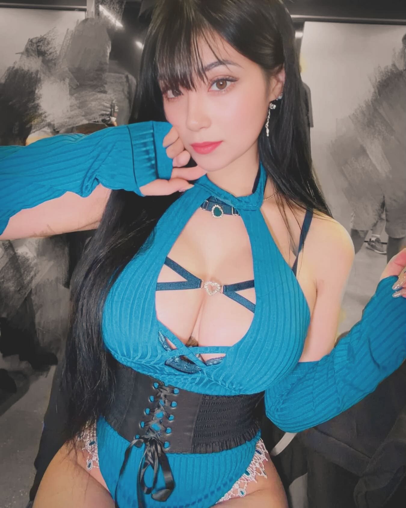 Against a grey backdrop, a woman with bangs poses in a teal ribbed halter top, detached sleeves, and a black lace-up corset belt.