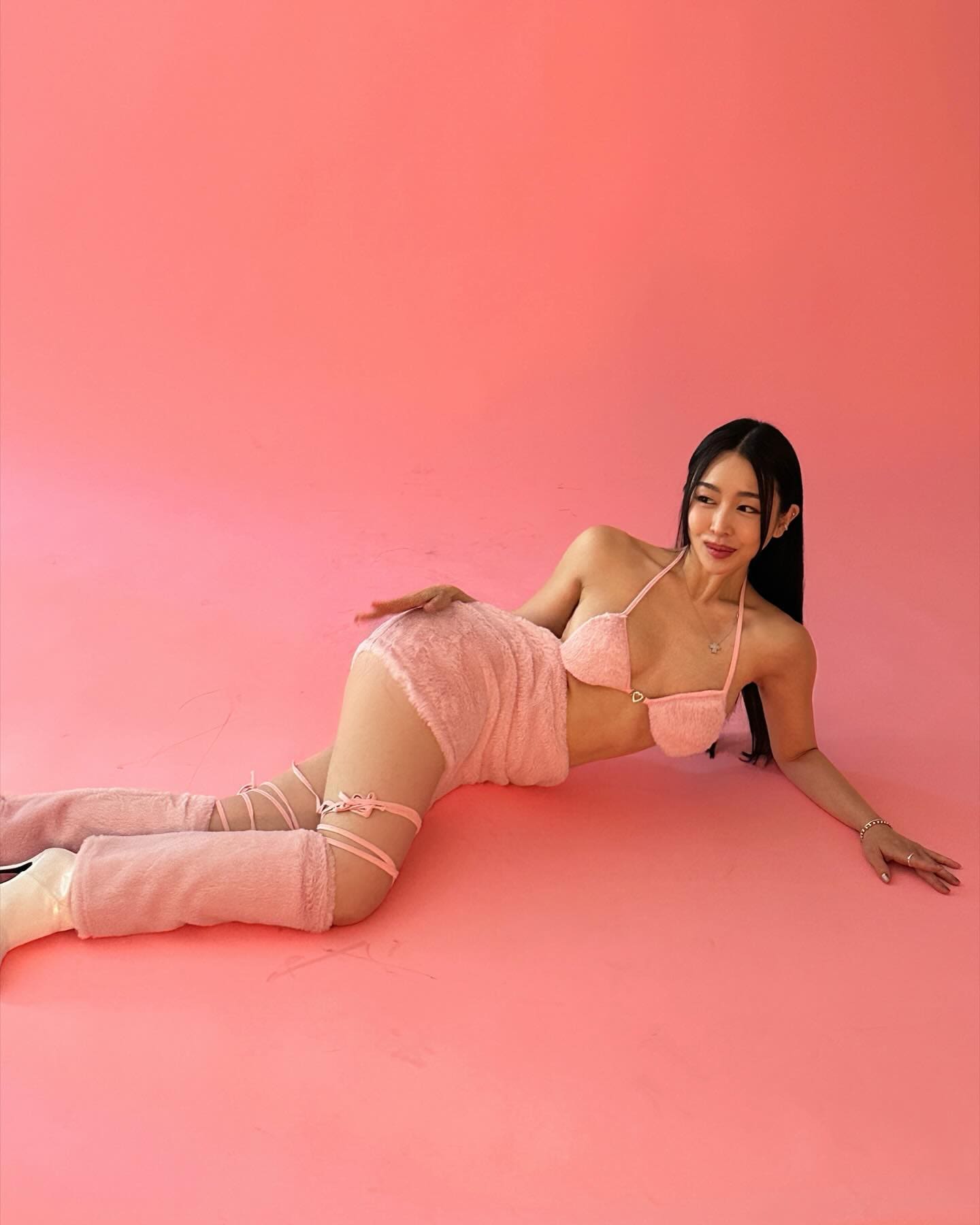 Reclining on a pink studio floor, Kali wears a fuzzy pink micro-bikini, mini skirt, and strappy thigh-high leg warmers.