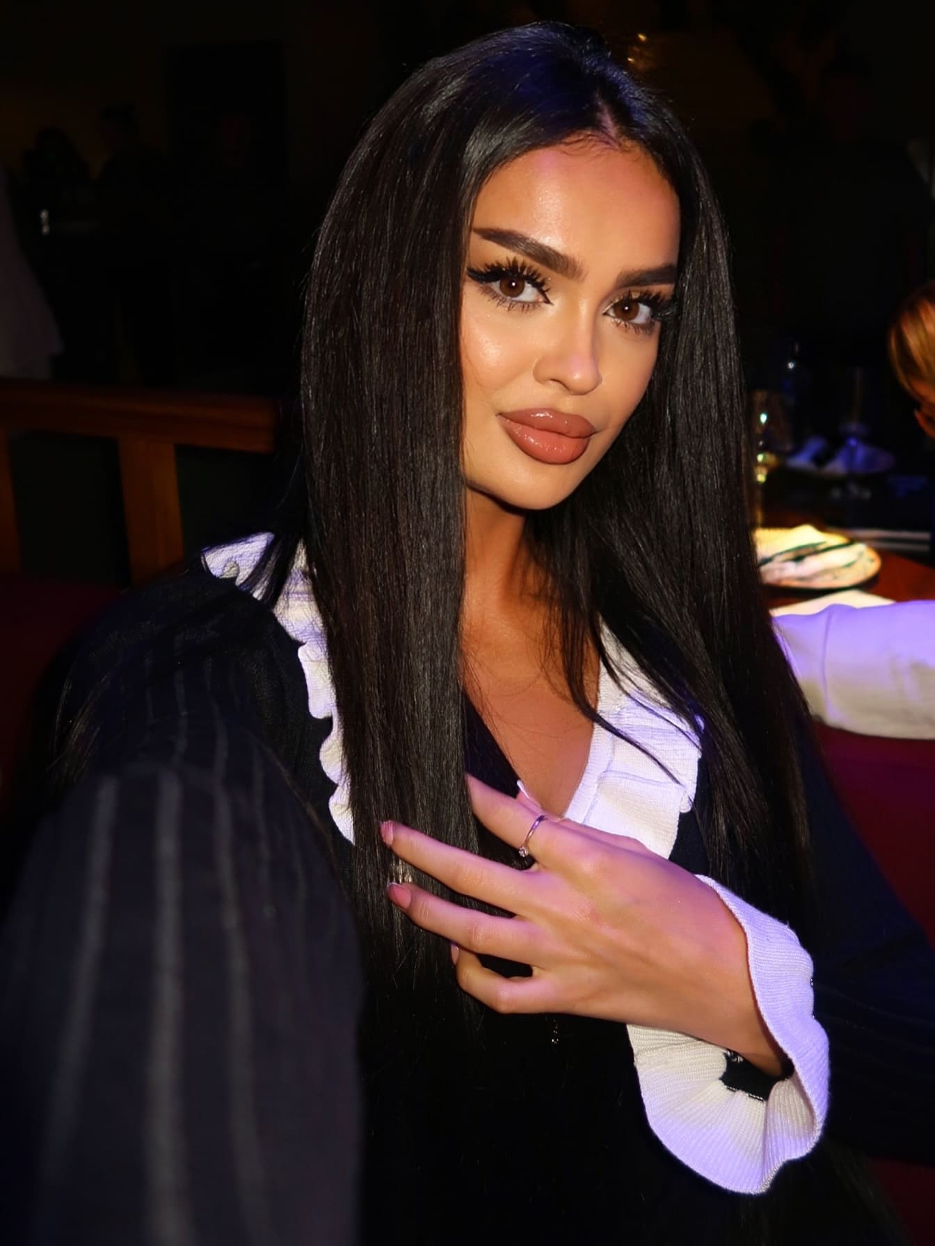 In a dim indoor setting, Sholpan wears a black pinstriped blazer over a white ruffled blouse, posing with a hand raised.