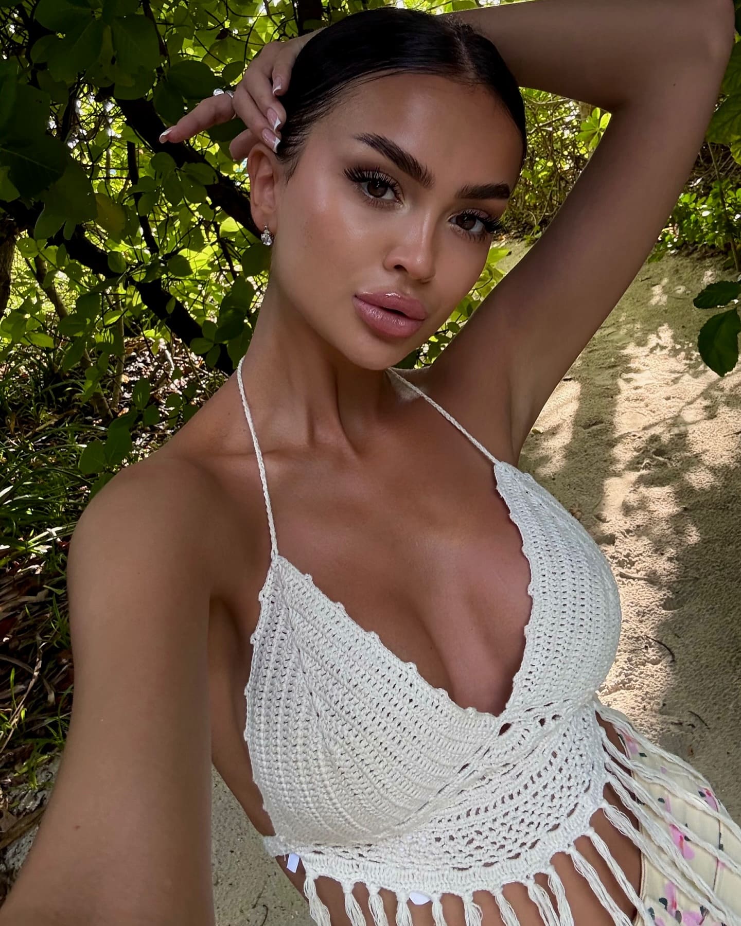 Posing outdoors with an arm behind her head, she wears a white crocheted halter top and floral bottom against green foliage.