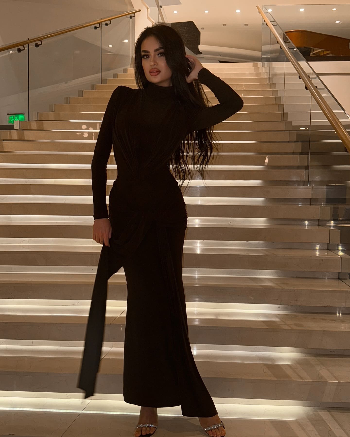 Standing on illuminated stairs, she wears a floor-length black dress with a thigh-high slit, posing with a hand near her head.
